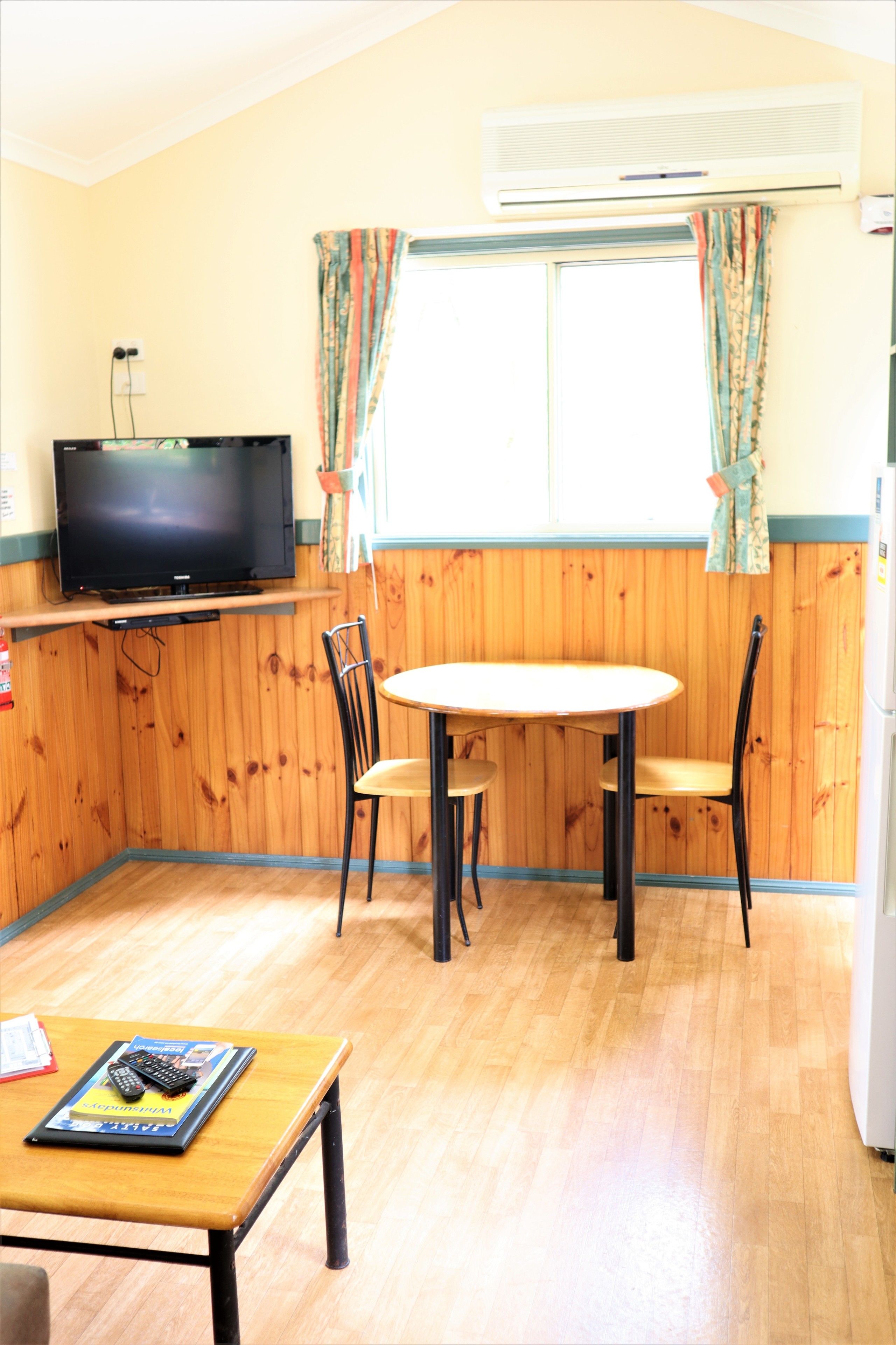 A room with a wooden paneled wall, a television on a shelf, a round table with two chairs, and a window with curtains.