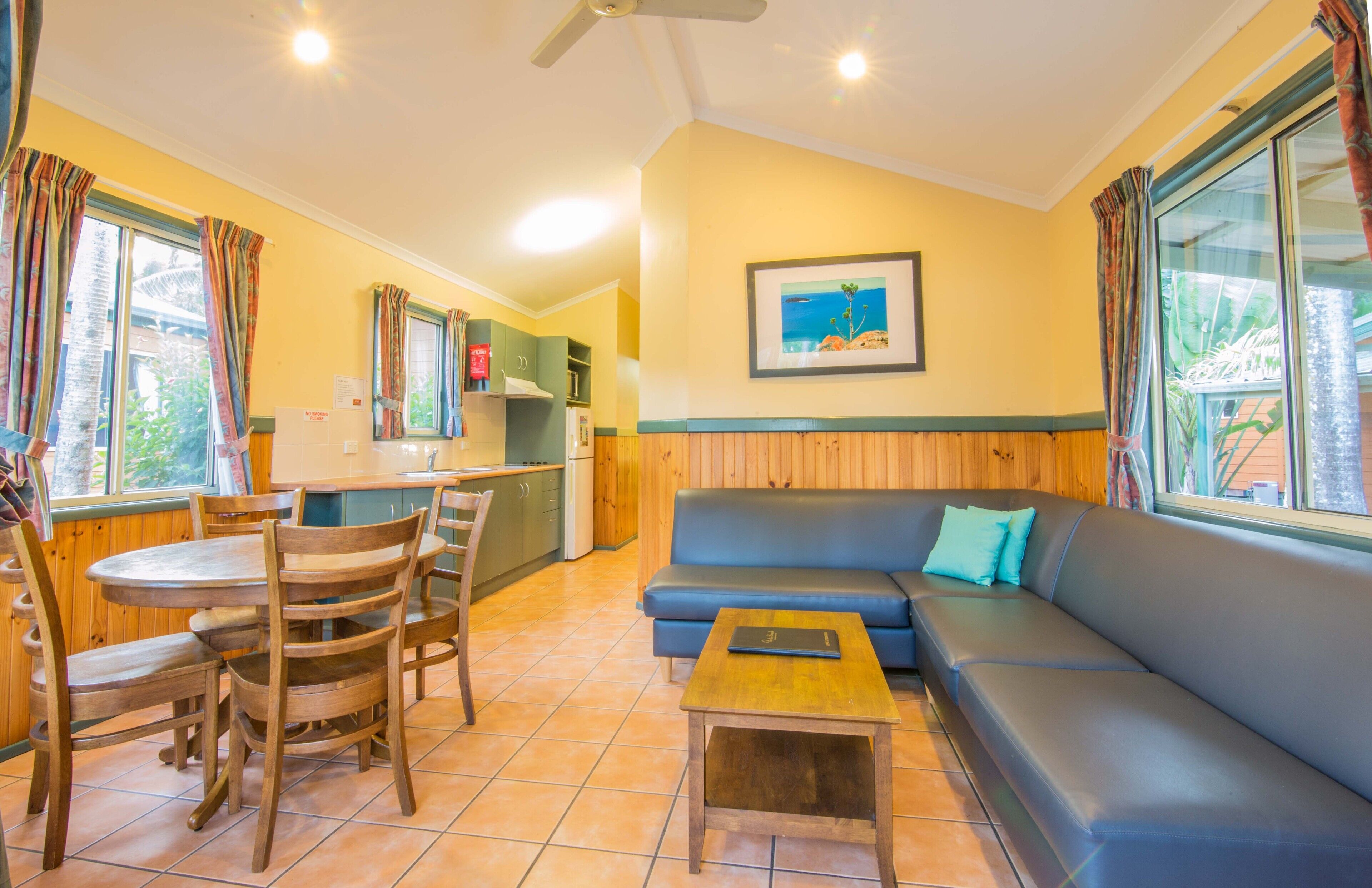 Una sala acogedora con un sofá azul, una mesa de comedor de madera y sillas. Se observa una zona de cocina con armarios y una ventana con cortinas.