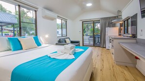 Une chambre d’hôtel avec un grand lit, un petit canapé, une kitchenette et un balcon offrant une vue sur les palmiers.