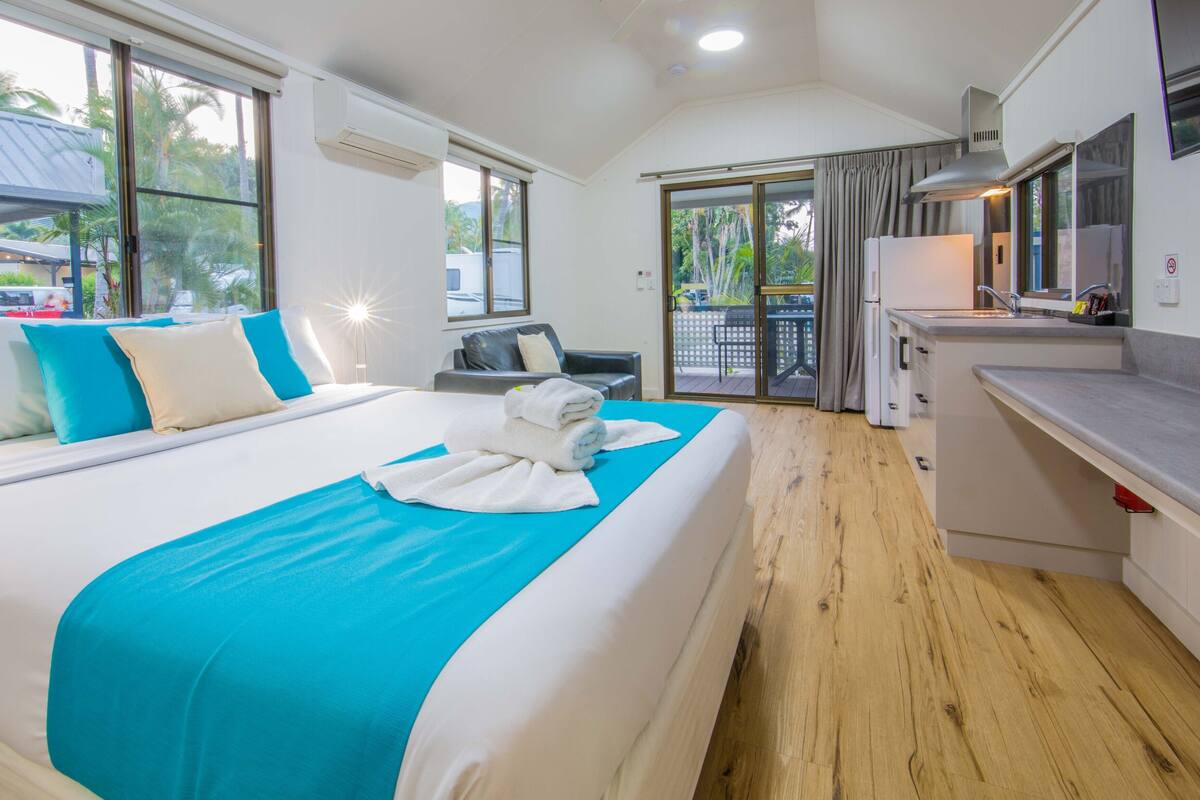 Une chambre d’hôtel avec un grand lit, un petit canapé, une kitchenette et un balcon offrant une vue sur les palmiers.