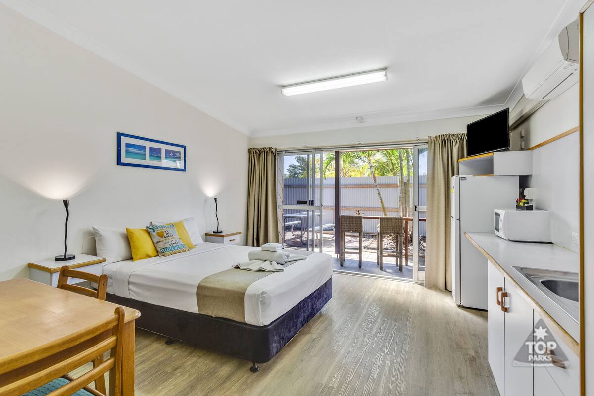 Une chambre d’hôtel comprenant un lit, un bureau, une chaise, une table, un coin cuisine et une porte-fenêtre coulissante donnant sur un espace extérieur.