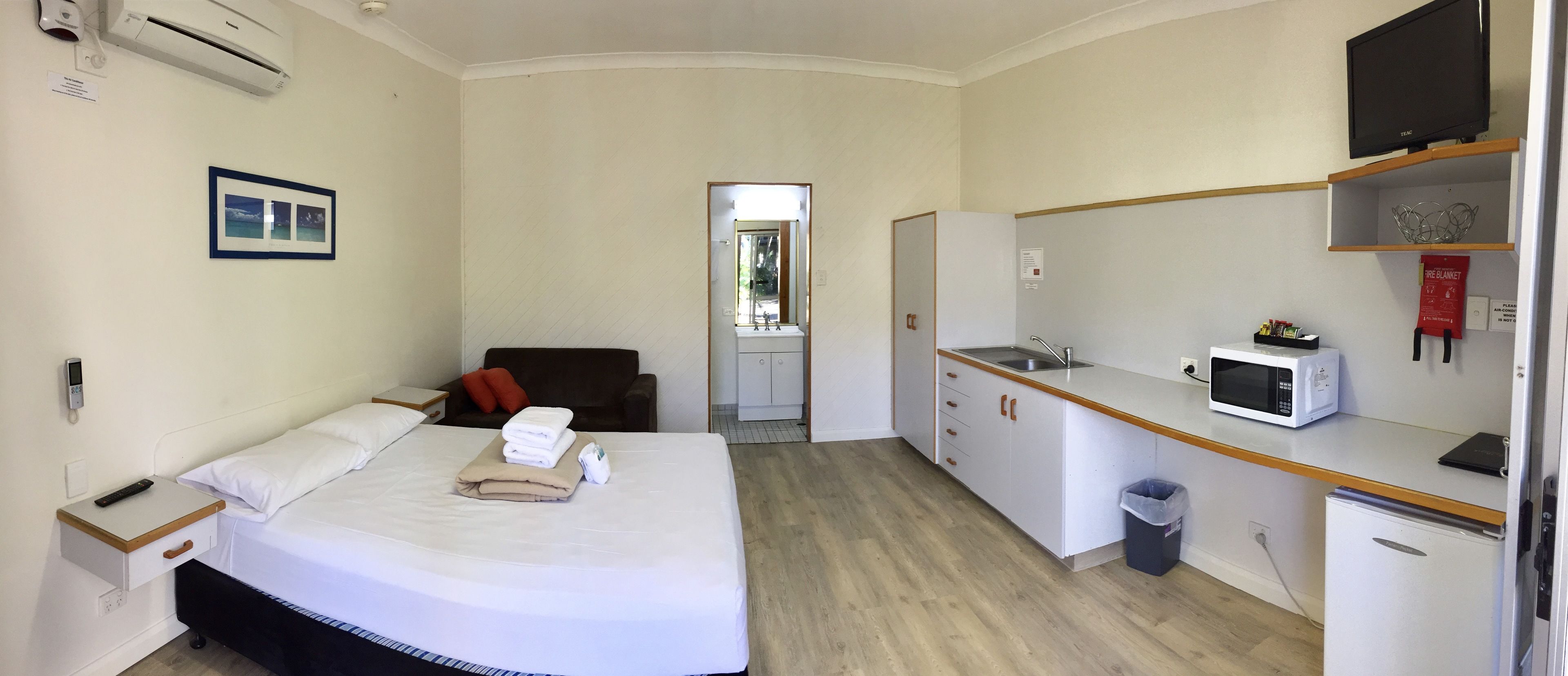 Una habitación de hotel con una cama, mesita de noche, un escritorio pequeño con microondas, lavamanos y un cesto de basura.