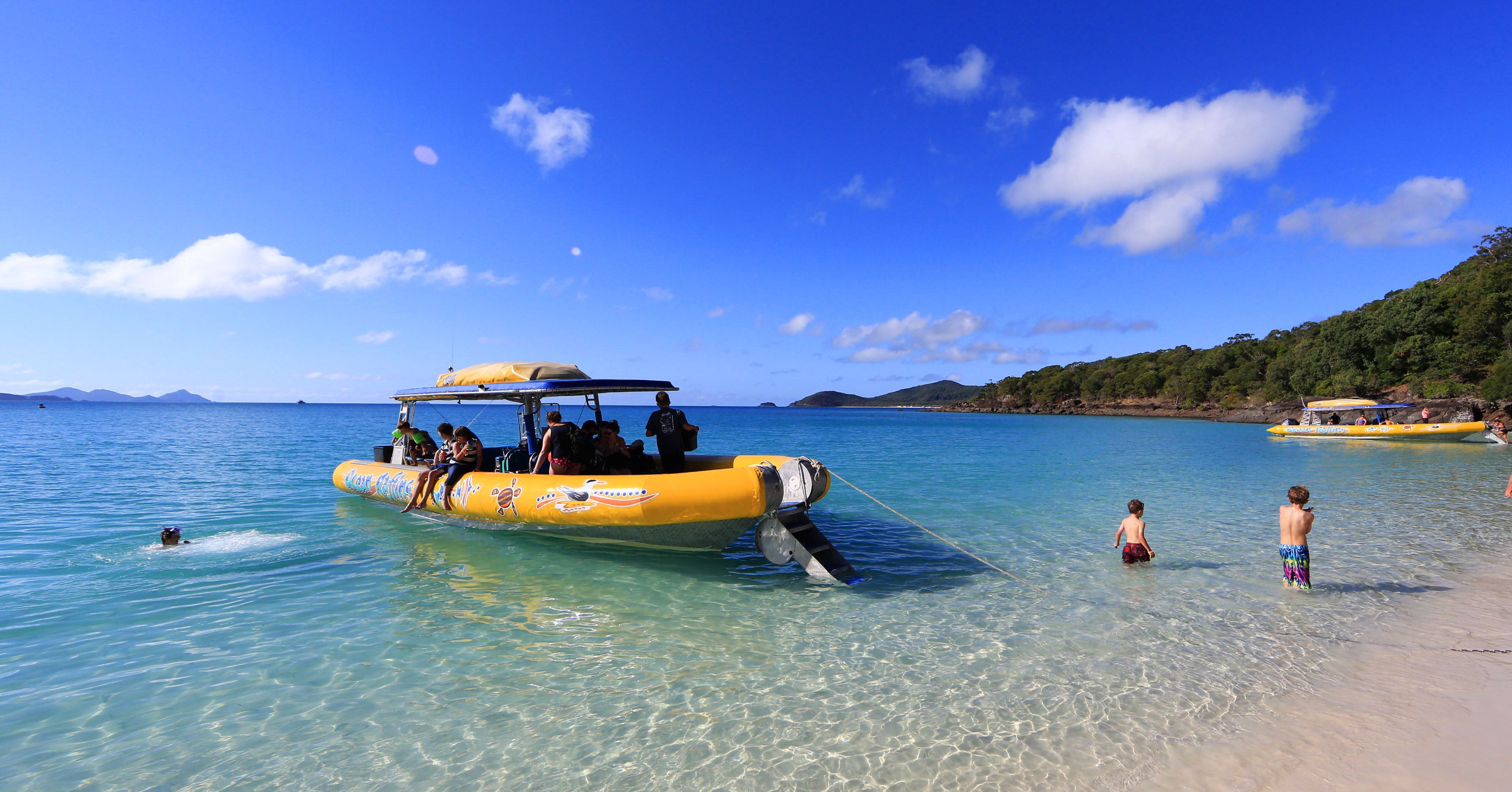 A yellow inflatable boat with people on it, floating on clear blue water near a beach.