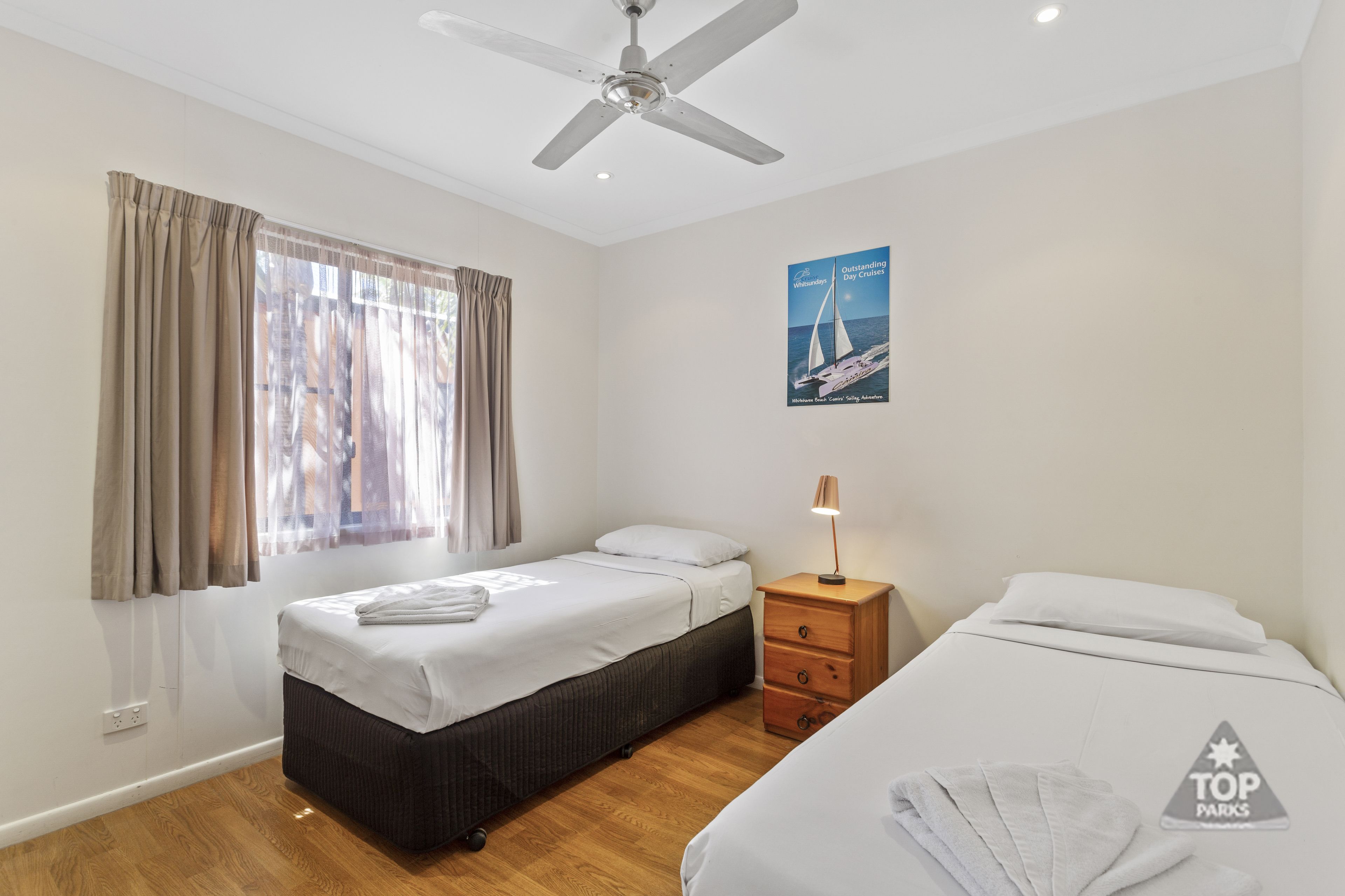A hotel room with two beds, a ceiling fan, a poster on the wall, and a wooden nightstand with a lamp.