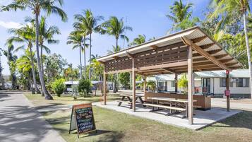 A covered picnic area with benches and picnic tables, surrounded by palm trees and a signboard.