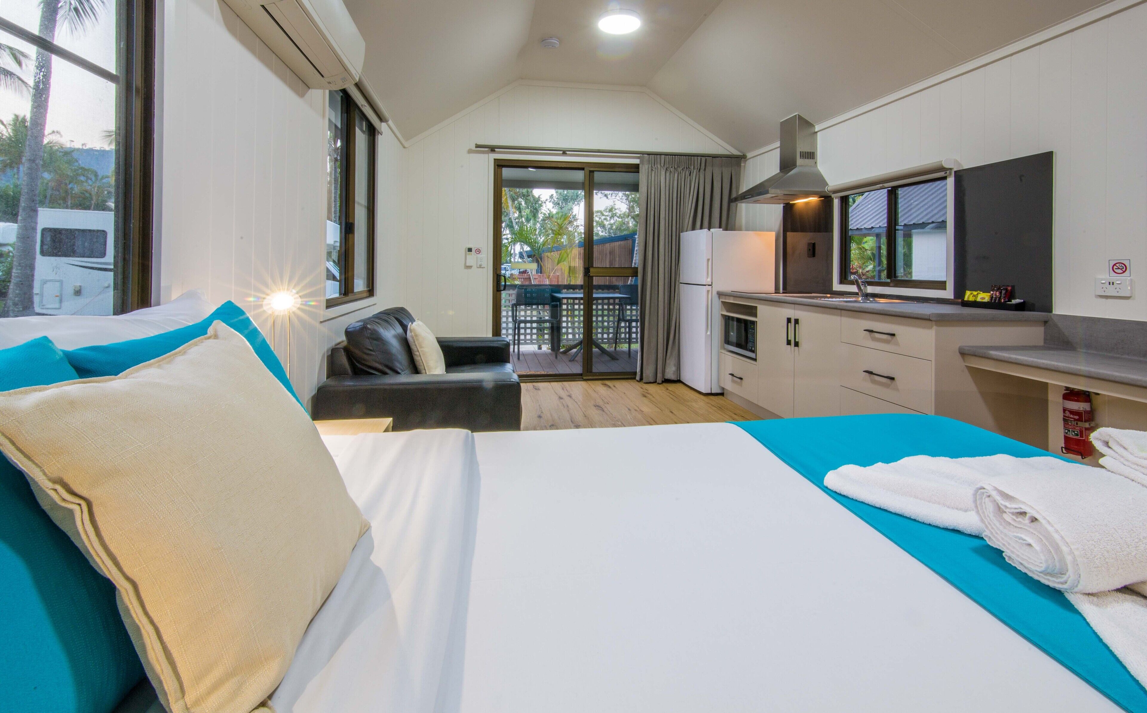 A modern hotel room with a bed, a sofa, a kitchenette, and a balcony with a view of palm trees.