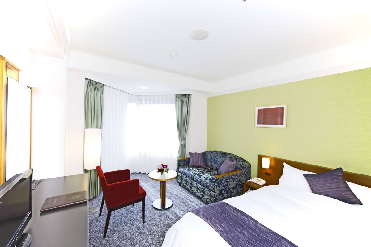 Una habitación de hotel con cama, sofá, escritorio con computadora y una mesita con un jarrón de flores.