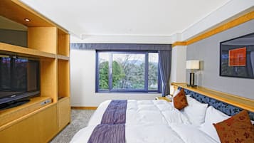 Habitación de hotel con una cama grande, un televisor sobre un mueble de madera, una ventana con vistas a árboles y una mesita de noche con una lámpara.