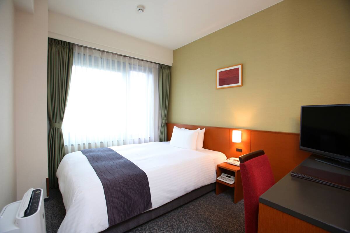 Habitación de hotel con una cama grande, un escritorio con televisor, una silla, una mesita de noche y una ventana con cortinas.