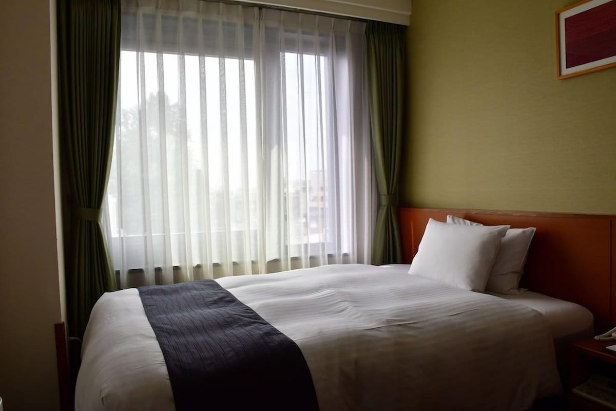 Une chambre d’hôtel avec un grand lit, une tête de lit en bois, une table de chevet et une fenêtre ornée de rideaux légers.