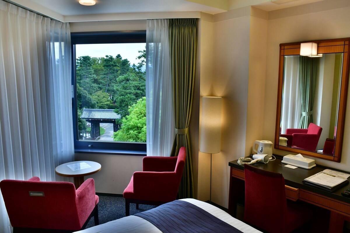 Una habitación de hotel con una cama, dos sillas rojas, una mesita redonda pequeña, un escritorio con lámpara, un espejo y una ventana con cortinas.