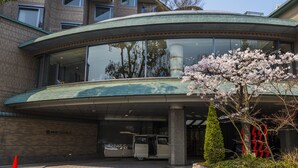 A modern building with a curved glass entrance, a vehicle parked outside, and a tree with pink blossoms. - Kyoto Garden Palace (Kyoto)