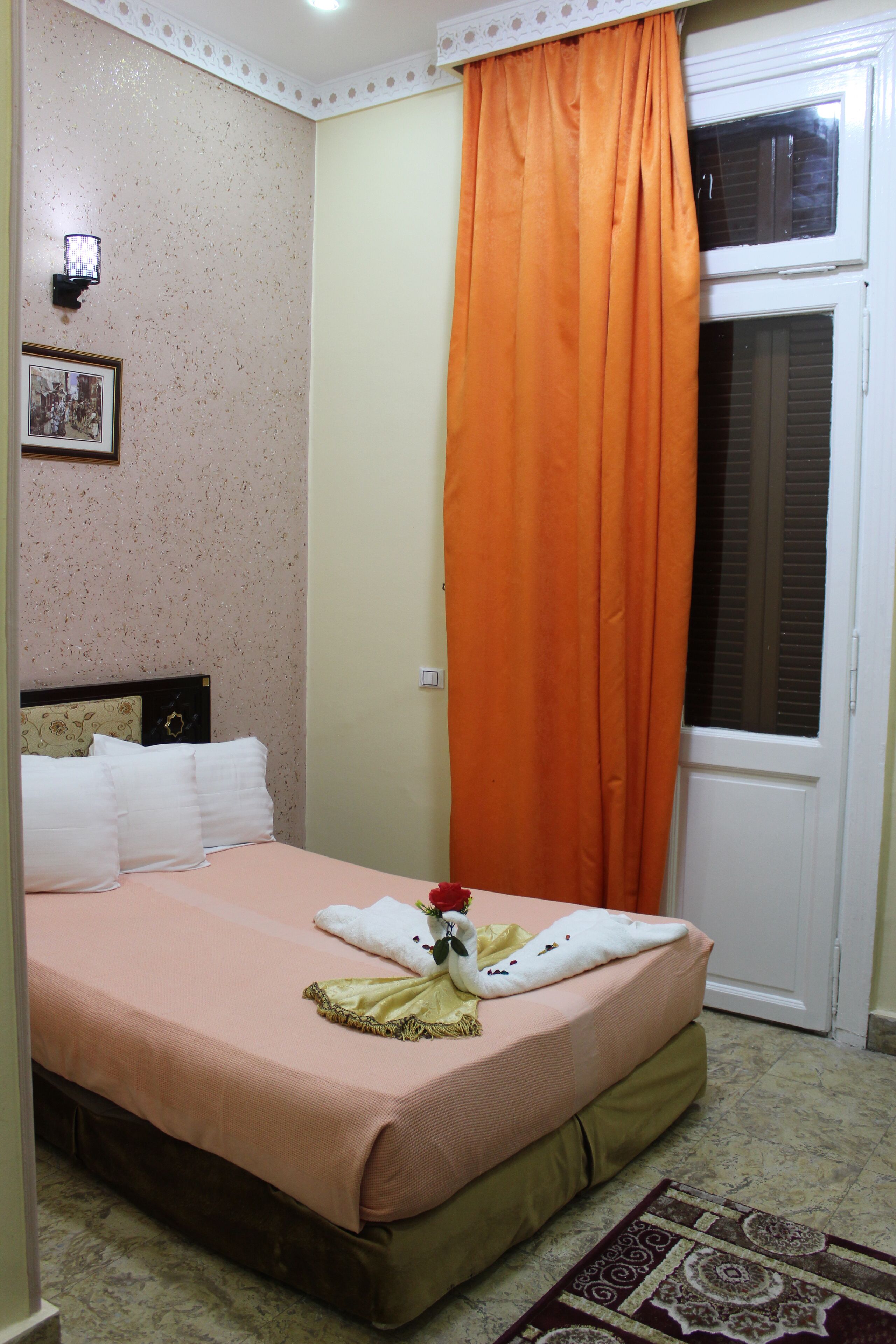 A bedroom with a bed, pillows, a nightstand, a window with blinds, and an orange curtain.