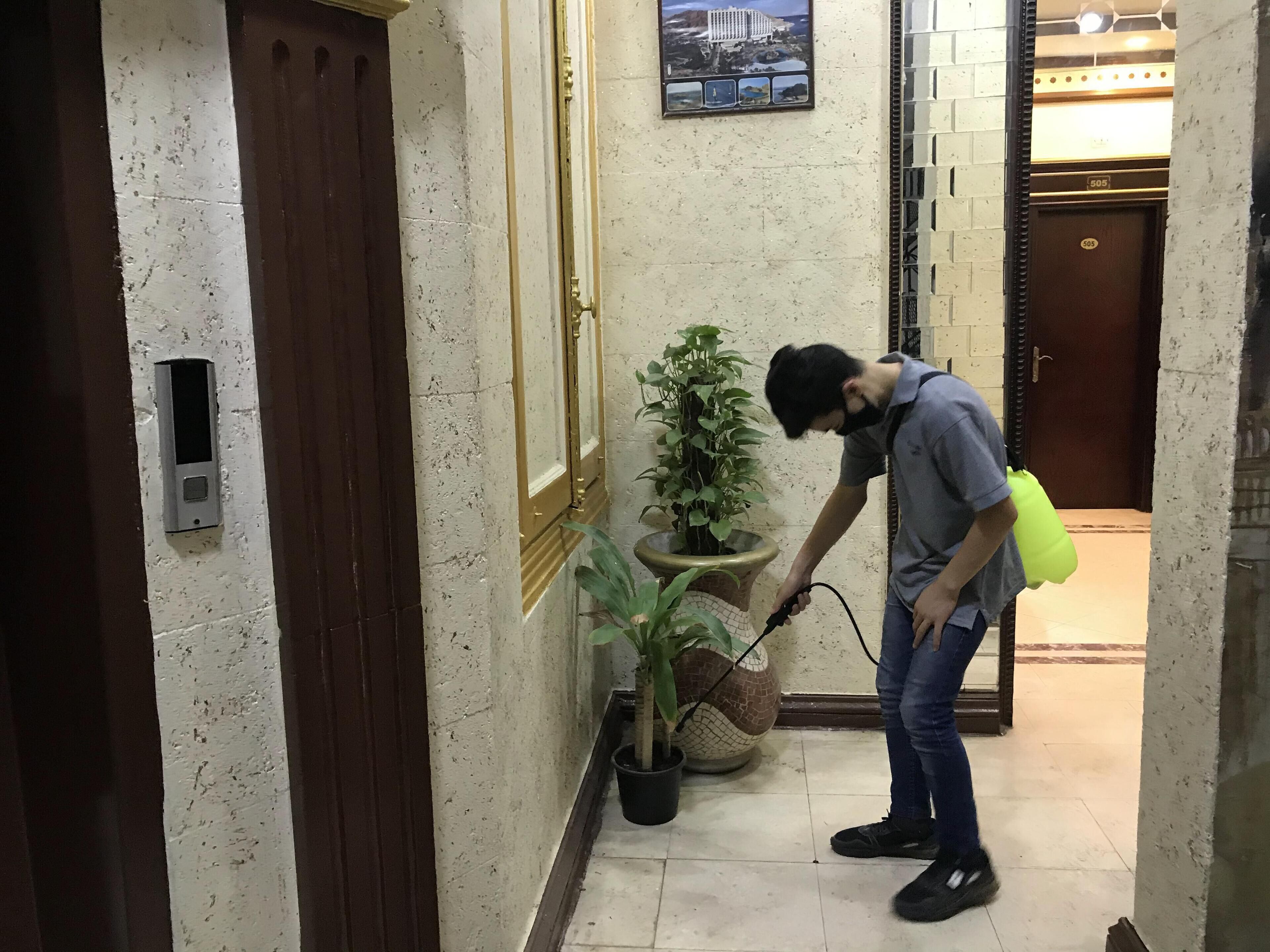 Un hombre limpiando un pasillo con un pulverizador. Hay una planta en maceta, una puerta con un número y un aplique de pared.