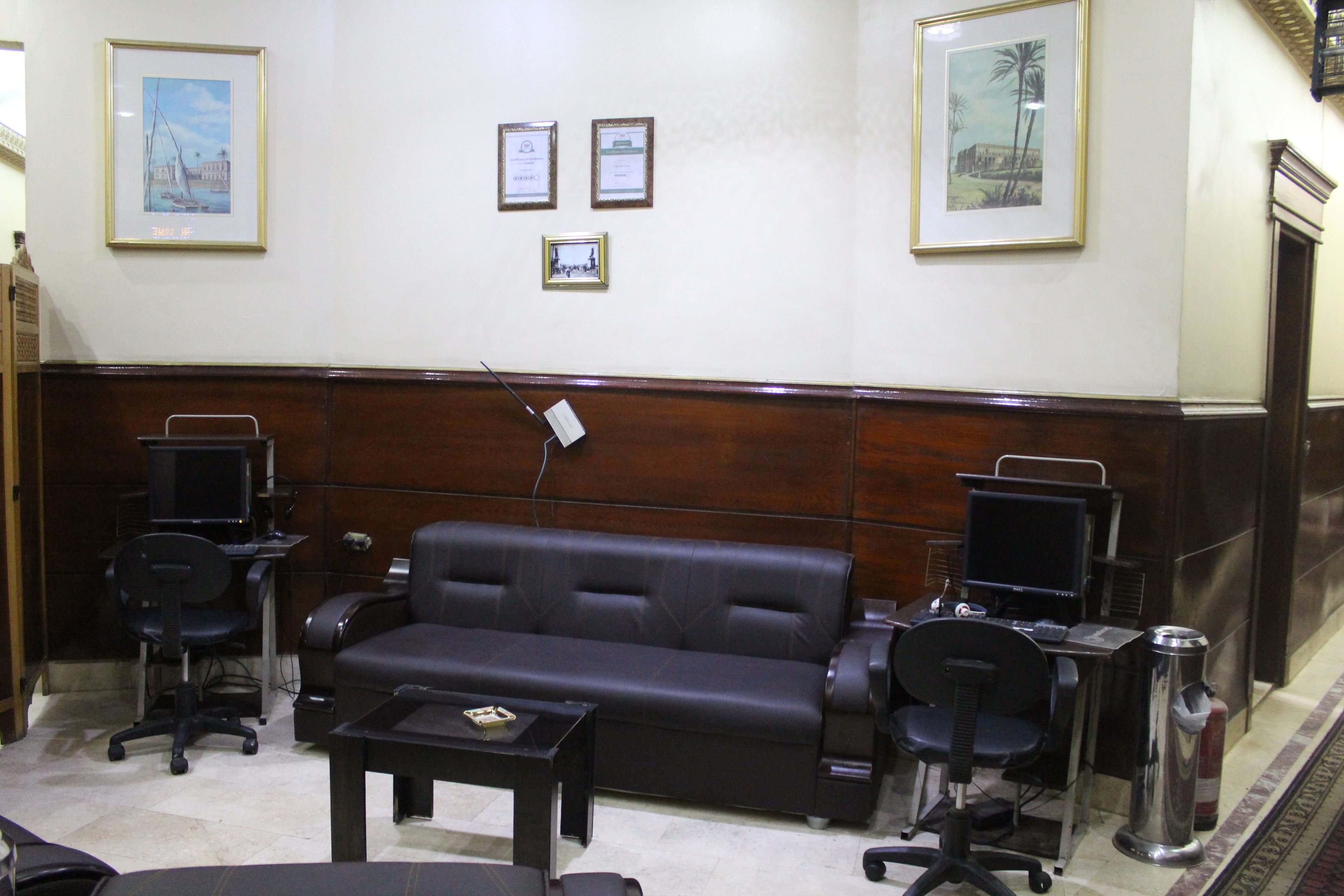 A conference room with a dark leather sofa, two office chairs, a small table, and two computer stations. There are framed certificates on the wall.