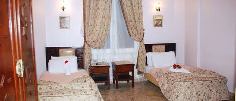 Una habitación con dos camas, una puerta de madera, una mesita y una ventana con cortinas.