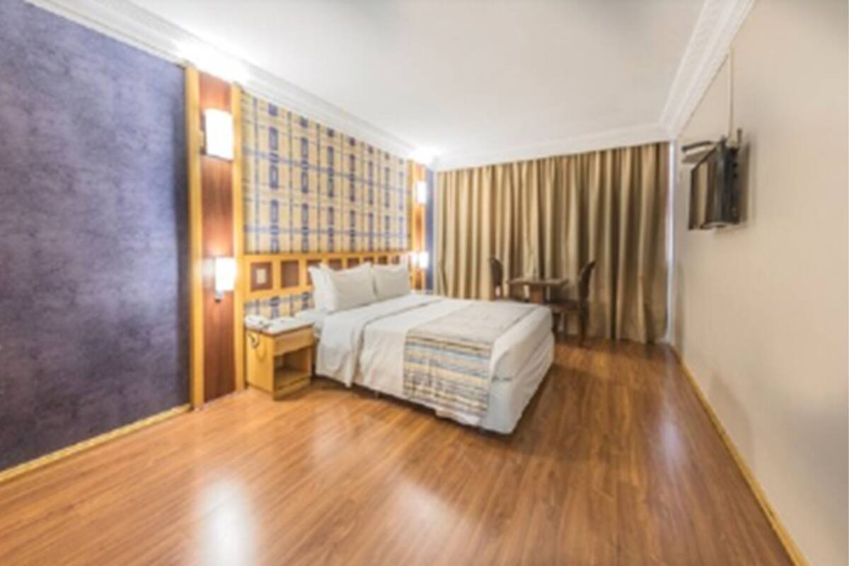 A hotel room with a wooden floor, a bed with a striped blanket, a nightstand, a wall-mounted TV, and a window with curtains.