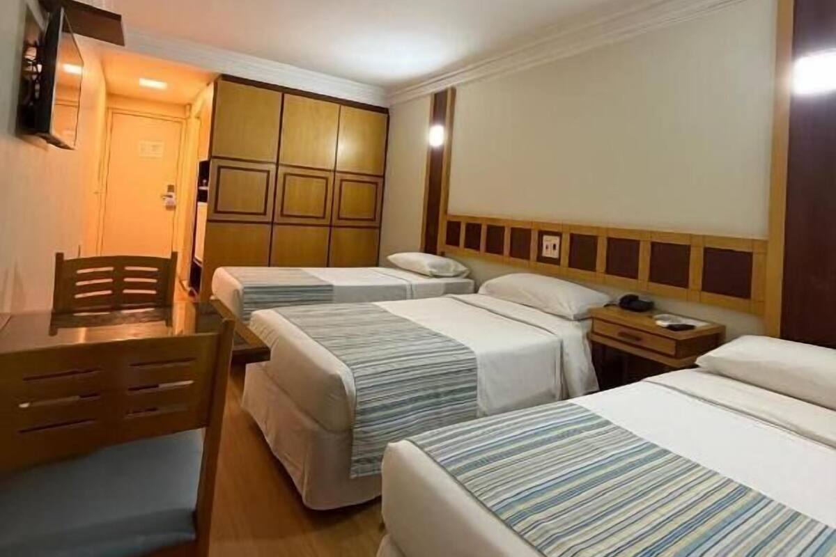 A hotel room with two beds, a wooden desk, a chair, a nightstand, and a wall-mounted TV.