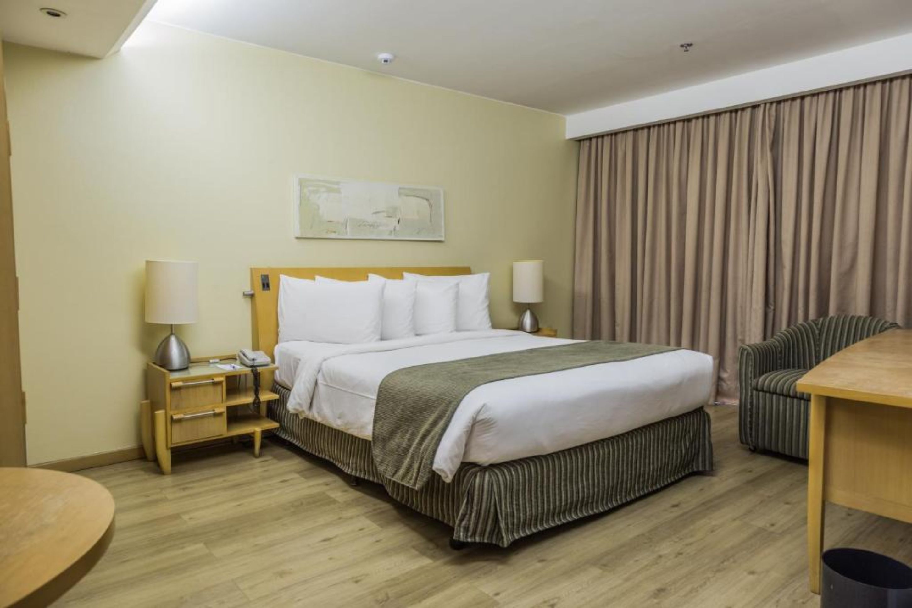 Quarto de hotel com cama, mesas de cabeceira, uma cadeira, uma mesinha e uma janela grande com cortinas.