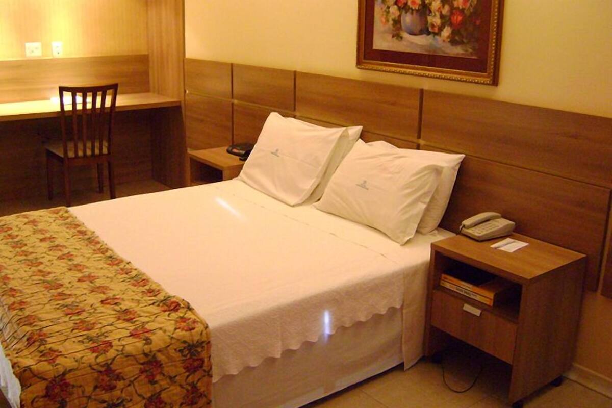 Une chambre d’hôtel avec un lit, un bureau, une chaise, une table de chevet avec un téléphone et un tableau au mur.