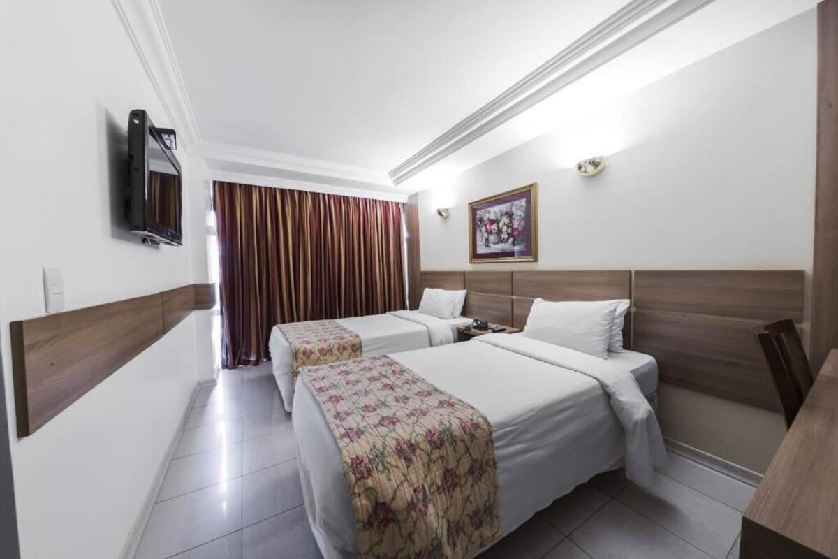 Une chambre d’hôtel avec deux lits, un téléviseur à écran plat fixé au mur, un tableau encadré et une fenêtre avec des rideaux.