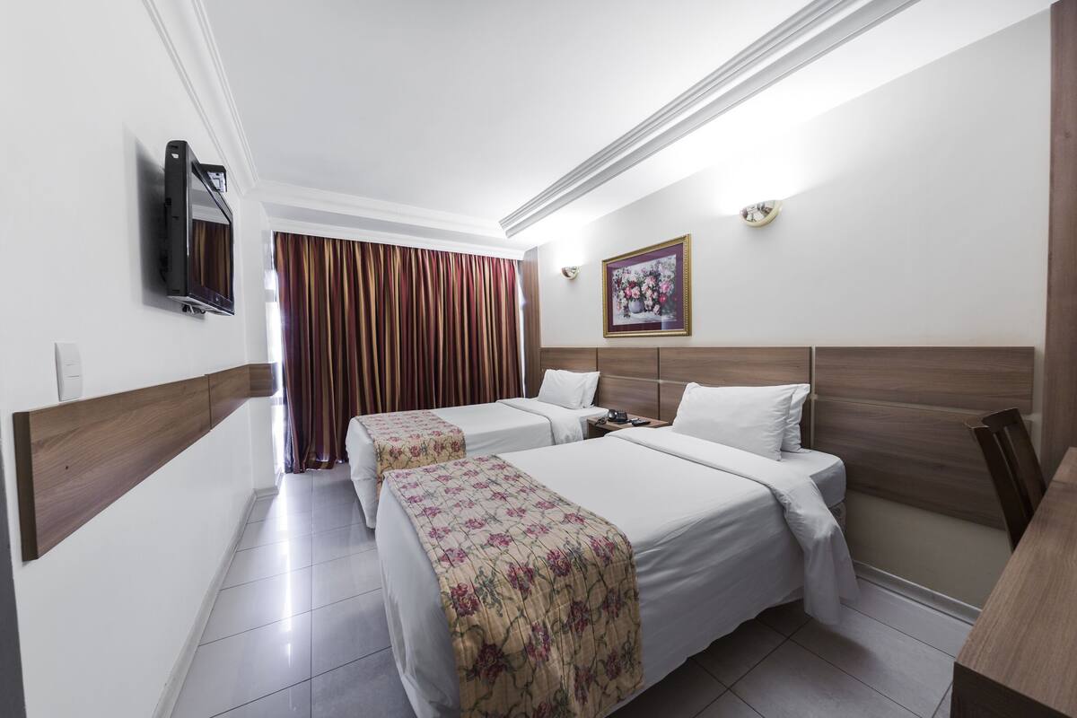 Une chambre d’hôtel avec deux lits, un téléviseur fixé au mur, un tableau et une fenêtre avec des rideaux.