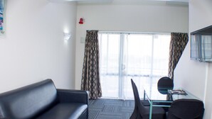 Una habitación de hotel con un sofá negro, una mesa de vidrio con una silla y un ventanal con cortinas estampadas.