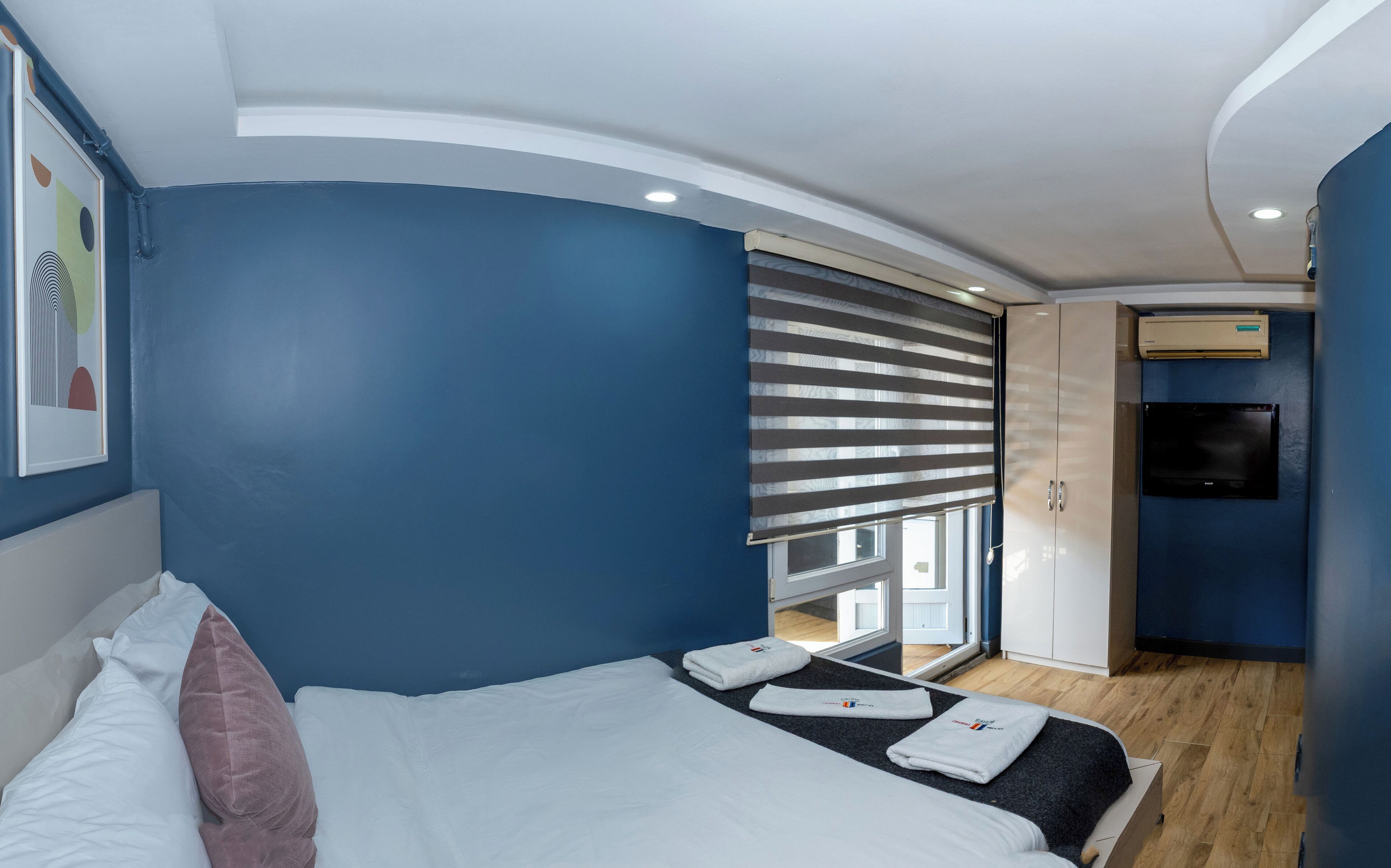 Quarto de hotel moderno com teto curvo, uma cama grande com almofadas brancas e cor-de-rosa, uma pequena mesa com toalhas, uma janela com persianas listradas e um guarda-roupa.