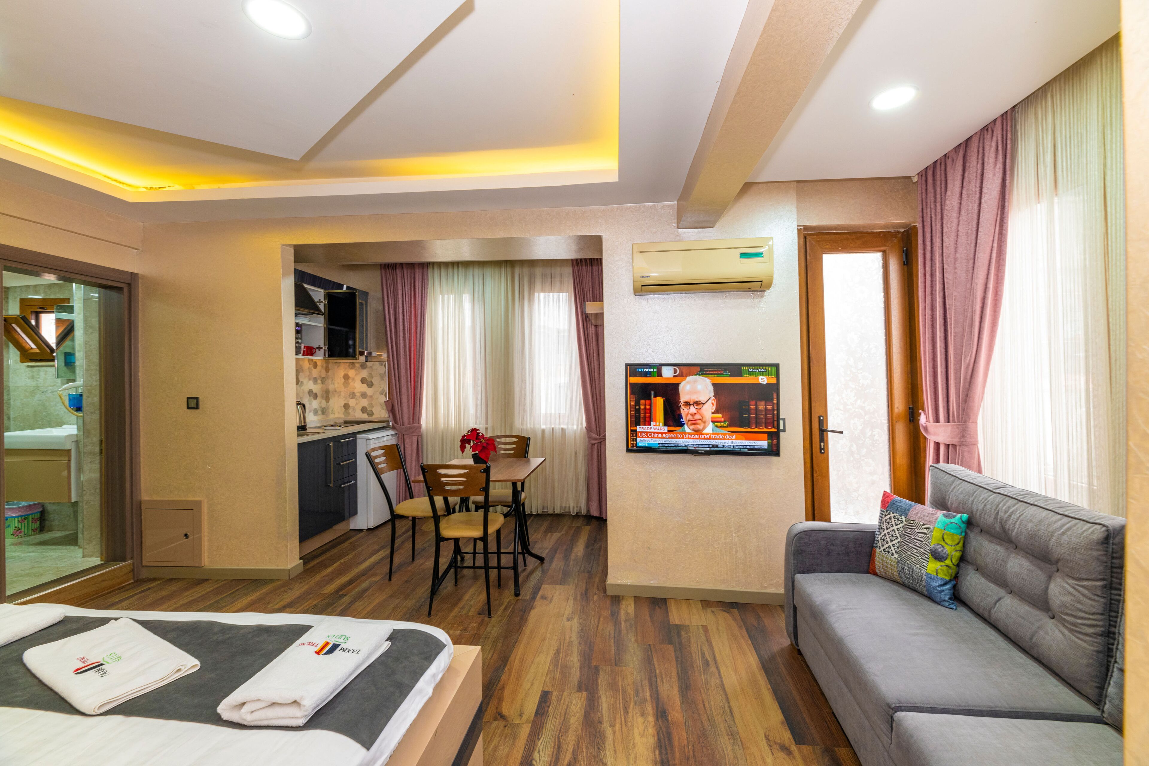 A hotel room with a bed, a sofa, a TV, a dining area, and an air conditioning unit.