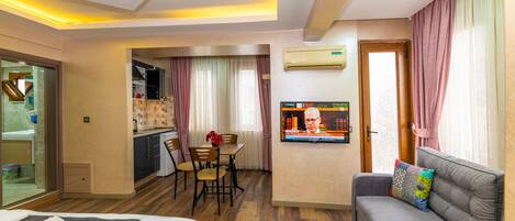A hotel room with a bed, a sofa, a TV, a dining area, and an air conditioning unit.