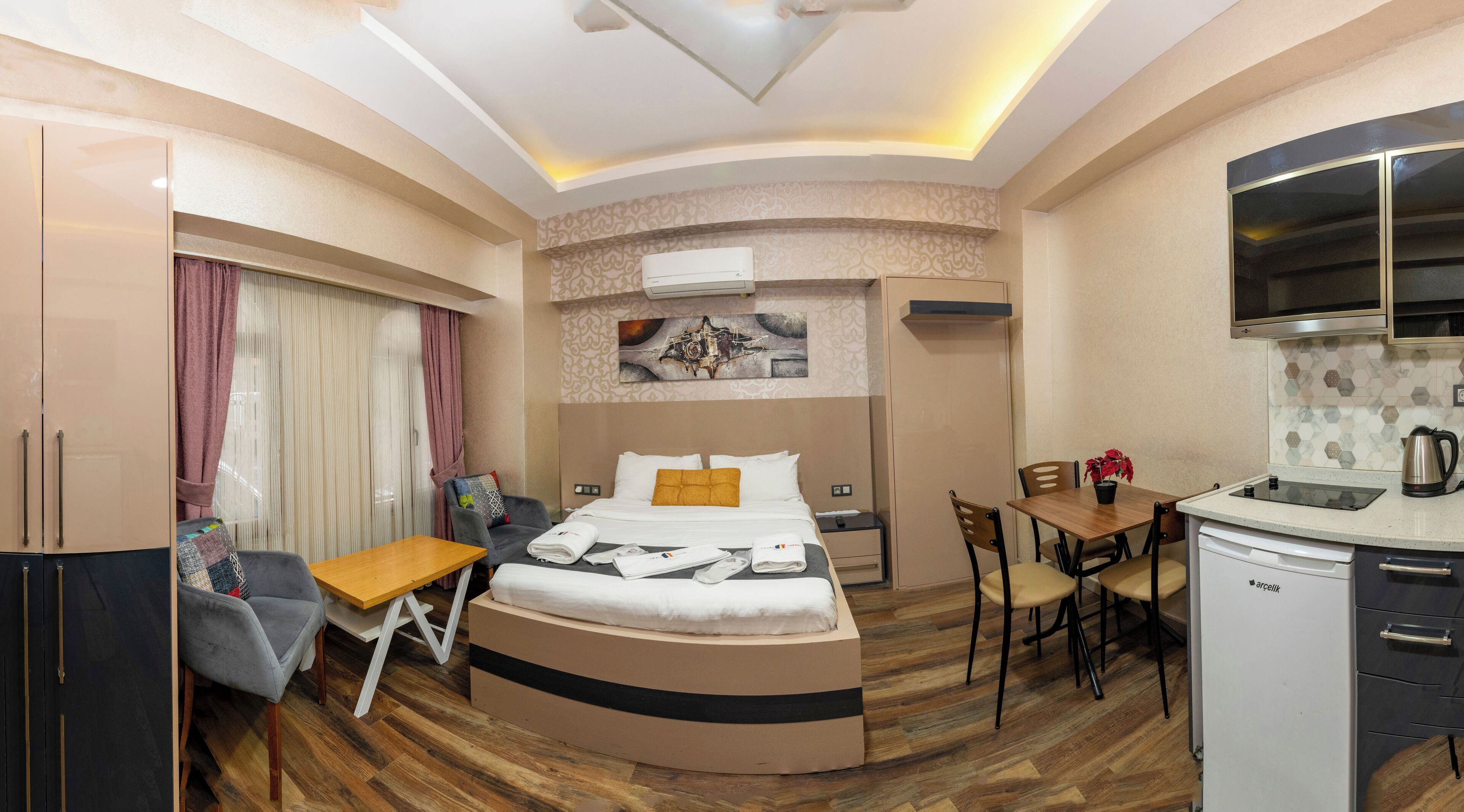 A compact hotel room with a bed, a small desk, a chair, a dining table with chairs, a kitchenette, and a large mirror.