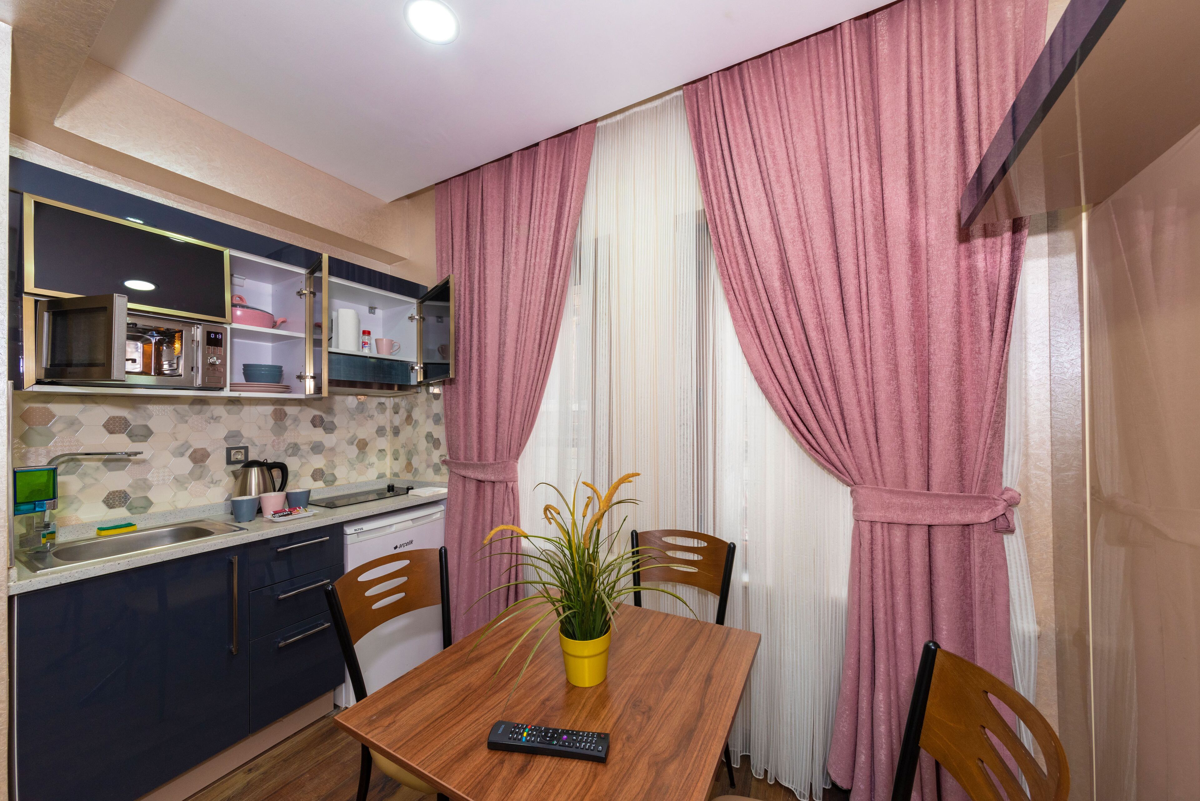 A compact kitchen with a built-in refrigerator, a microwave, and a sink. There is a dining table with chairs and a potted plant.