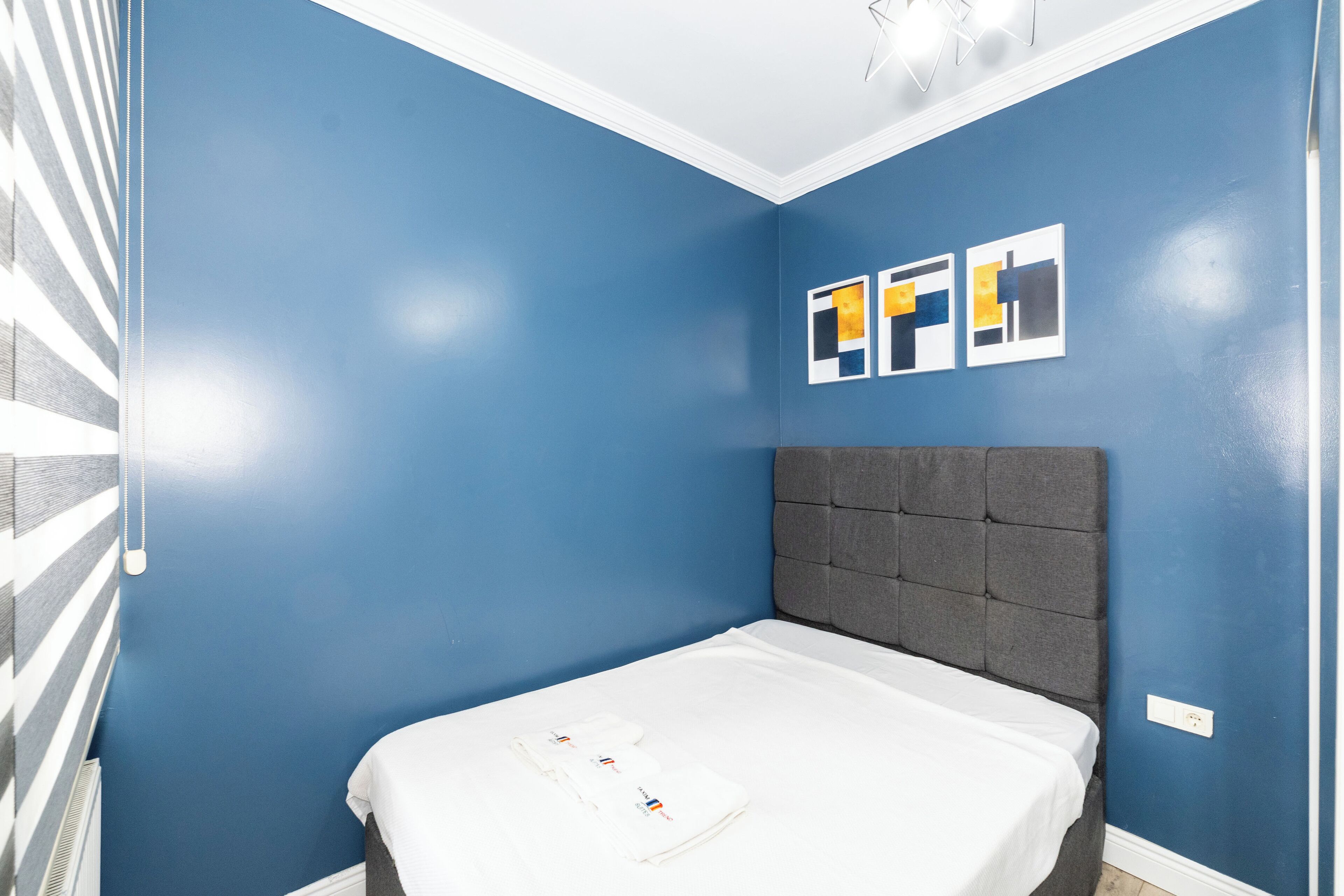 Ein Schlafzimmer mit einem Einzelbett, blauen Wänden und einem grauen Kopfteil. An der Wand hängen drei gerahmte Kunstwerke.