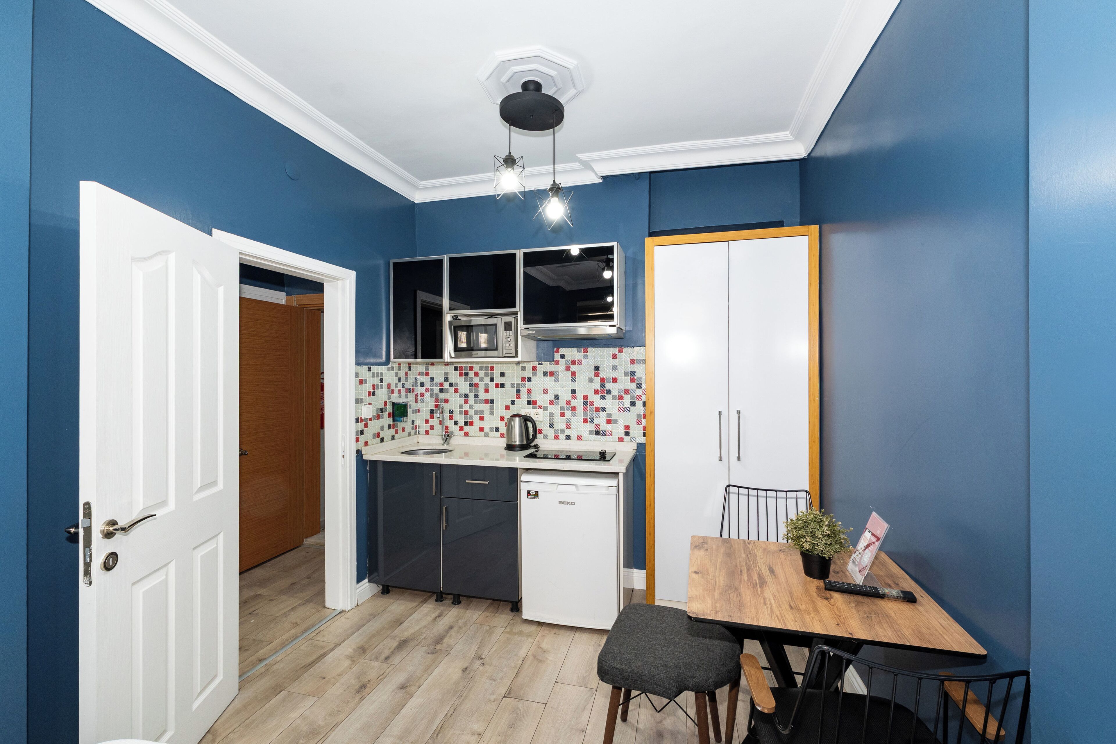 A compact kitchen with a microwave, sink, and a small refrigerator. There's a wooden dining table with a chair and a potted plant.