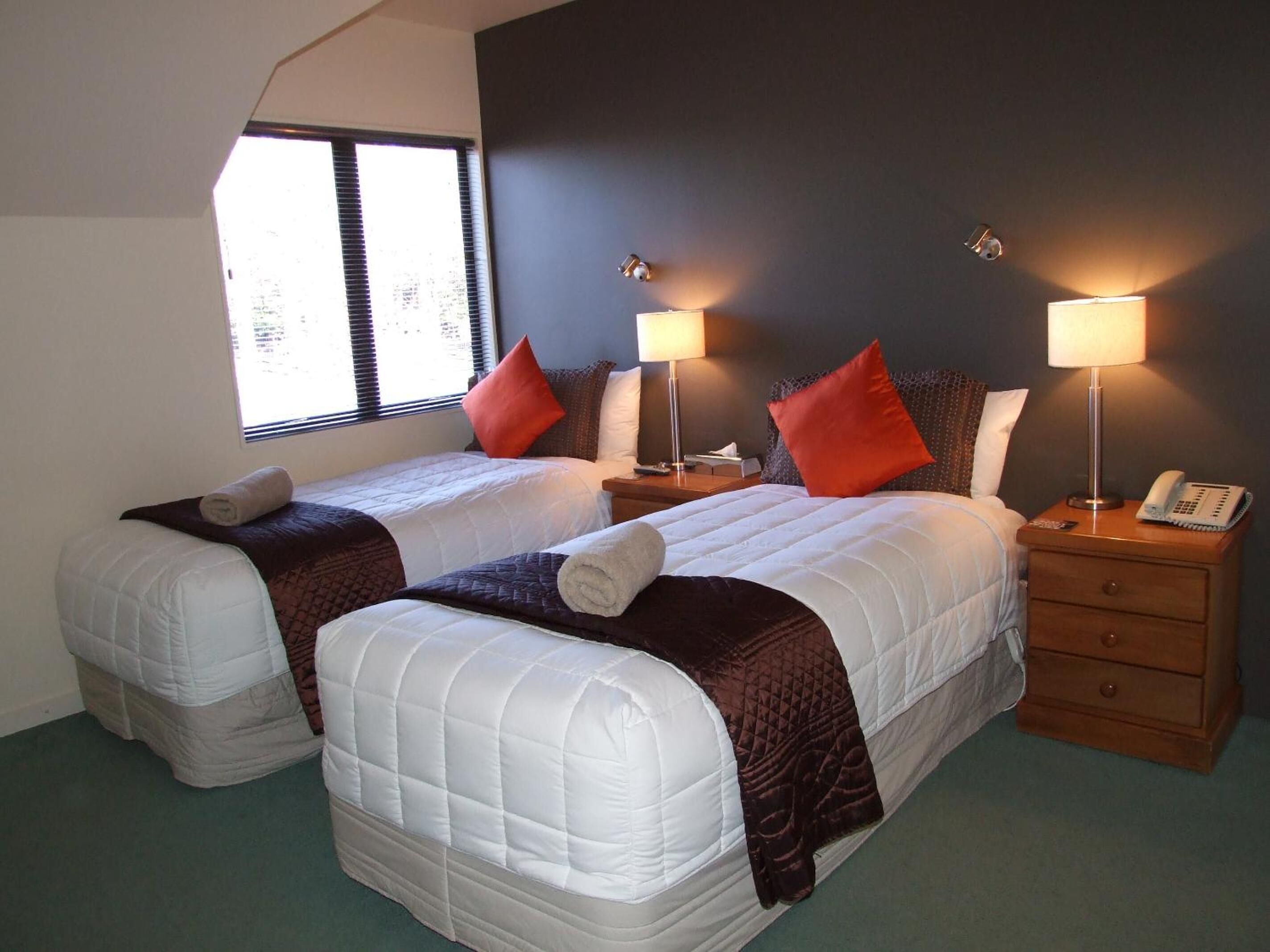 A hotel room with two beds, a nightstand with a lamp, a telephone, and a window with blinds.