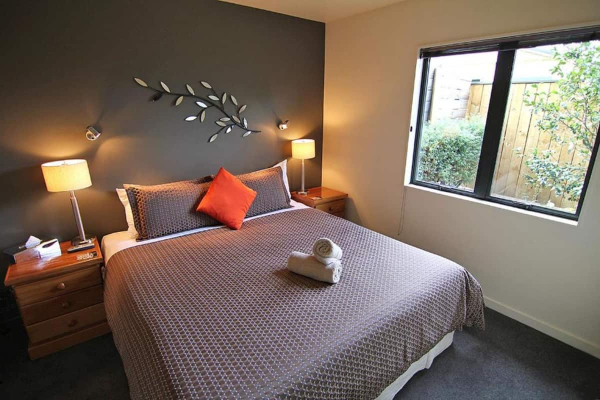 Un lit bien fait, recouvert d’une couvre-lit à motifs, deux lampes de chevet, un élément décoratif mural et une fenêtre donnant sur un paysage verdoyant.
