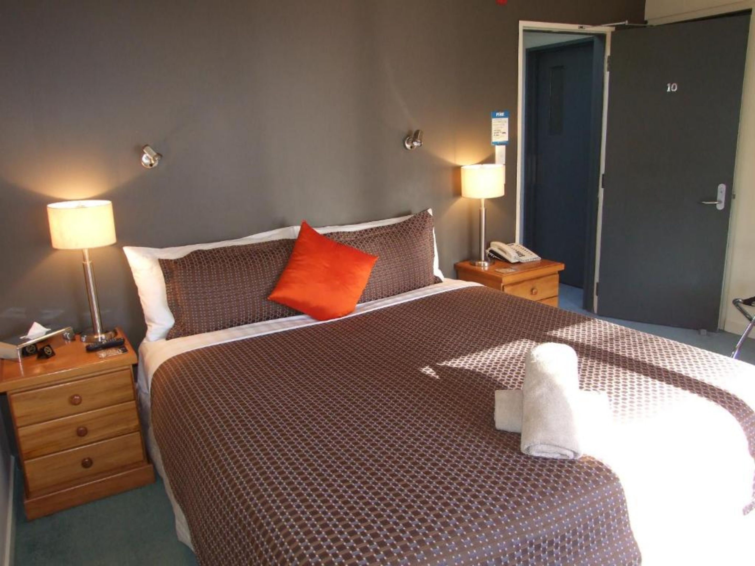 Ett hotellrum med en säng, två sängbord, ett litet bord, en lampa, en telefon och en dörr.