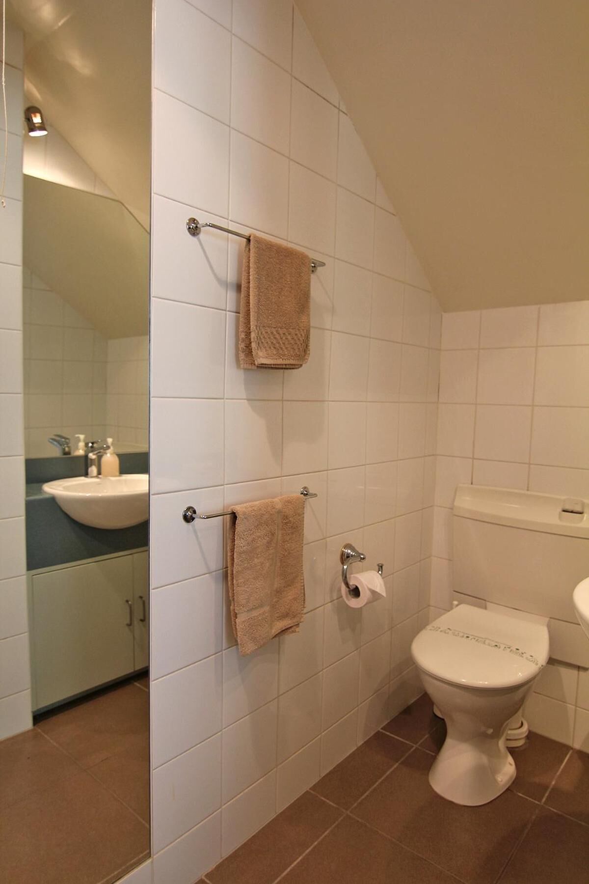 Une salle de bain avec des murs recouverts de carreaux blancs, un lavabo, des toilettes et deux serviettes suspendues à des porte-serviettes.