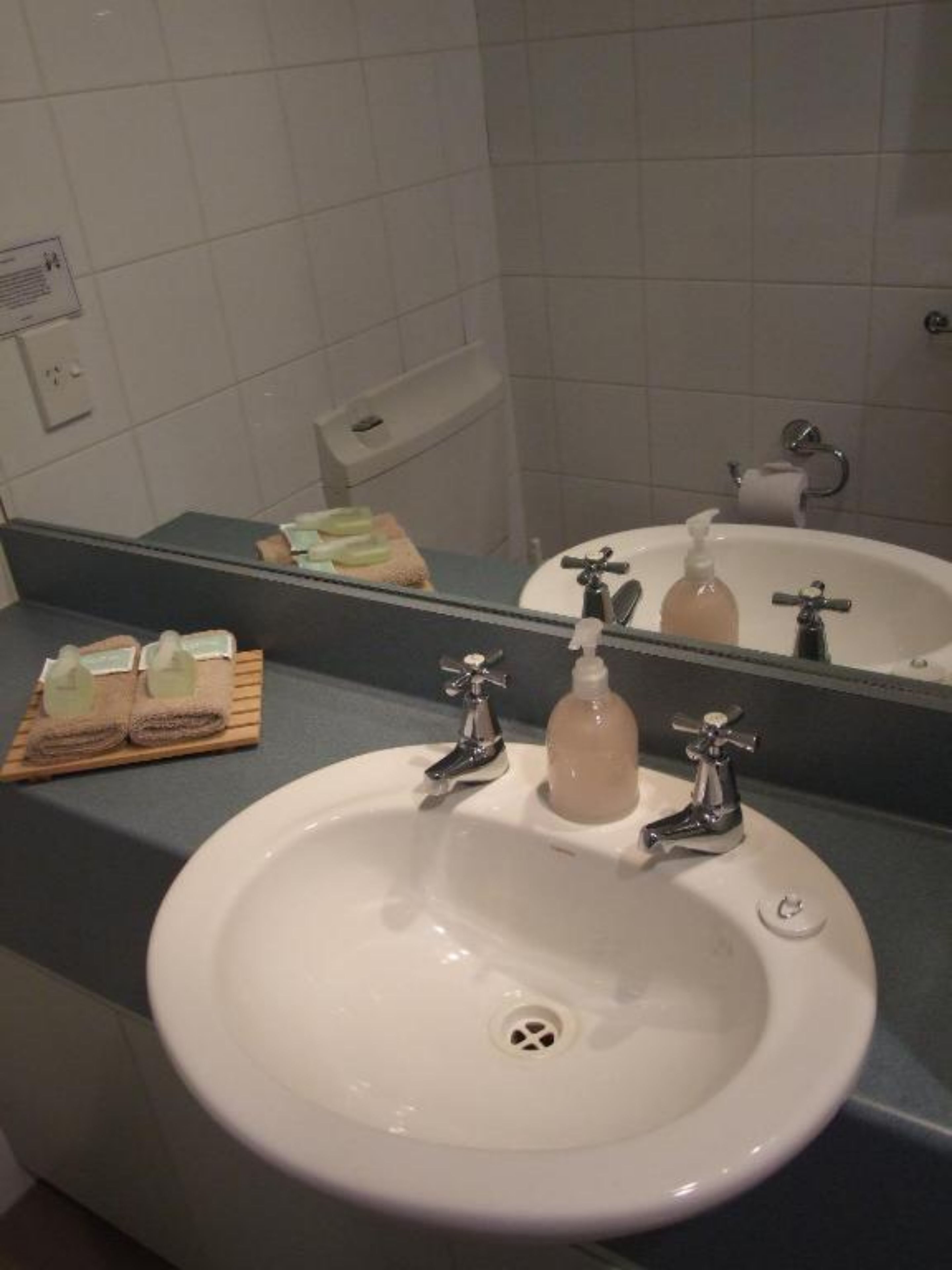 A bathroom with a white sink, two faucets, a soap dispenser, and a mirror reflecting a toilet paper holder and a towel dispenser.