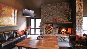 Een gezellige woonkamer met een stenen openhaard, leren banken, een houten salontafel en een flatscreen televisie die boven de openhaard is bevestigd.