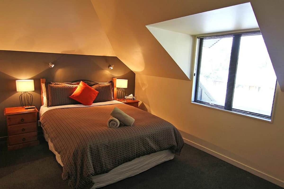 Une chambre avec un plafond en pente, une grande fenêtre avec des stores, un lit avec une serviette roulée, deux lampes de chevet et une table de chevet avec une lampe.