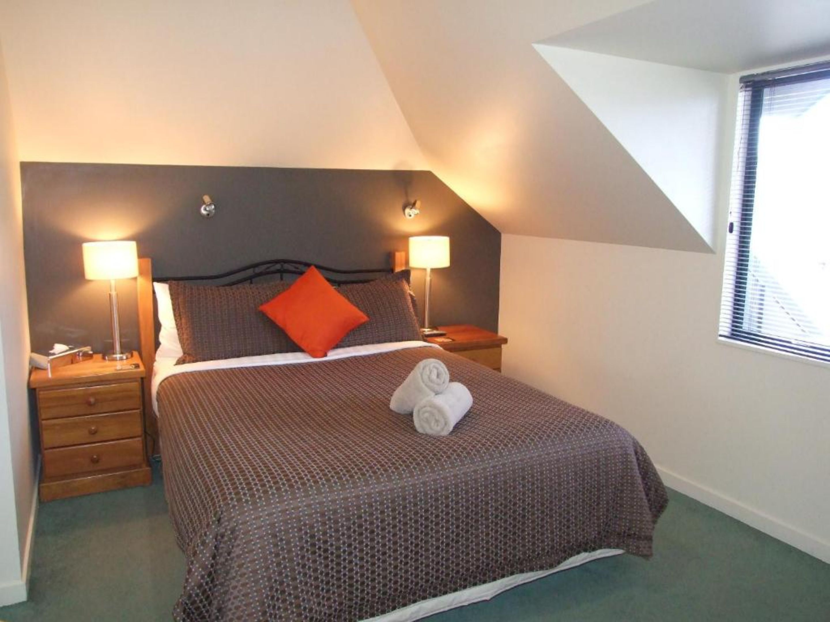 A neatly made bed with a brown patterned bedspread, two pillows, and a rolled towel. There are two bedside tables with lamps, a nightstand, and a window with blinds.