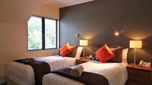 Een hotelkamer met twee bedden, elk voorzien van een kussen en dekbed, een nachtkastje met een lamp en telefoon, en een raam met jaloezieën.
