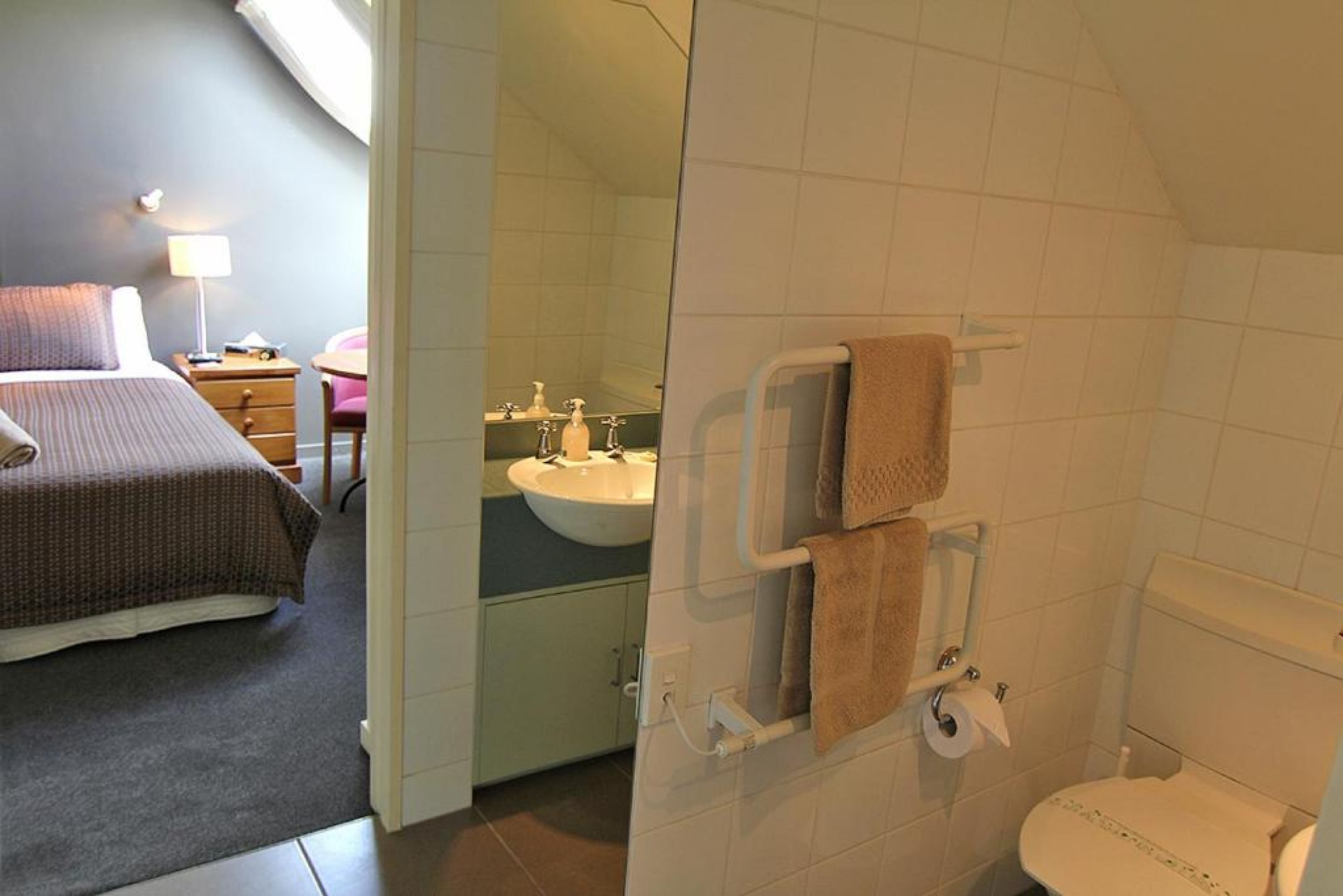 A hotel room with a bed, a bedside table with a lamp, a bathroom with a sink, mirror, and toilet.