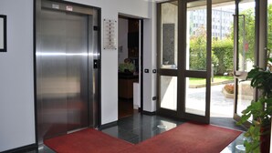 An interior view of a hotel lobby with an elevator, a reception area, and a glass door leading outside. - Residence Le Corti (Milan)