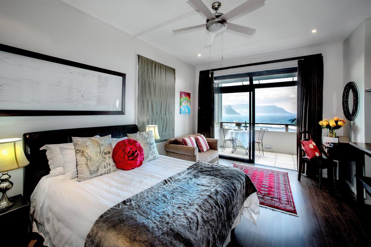 Quarto de hotel moderno com uma cama grande, um ventilador de teto, vista para o mar e uma varanda com mesa e cadeiras.