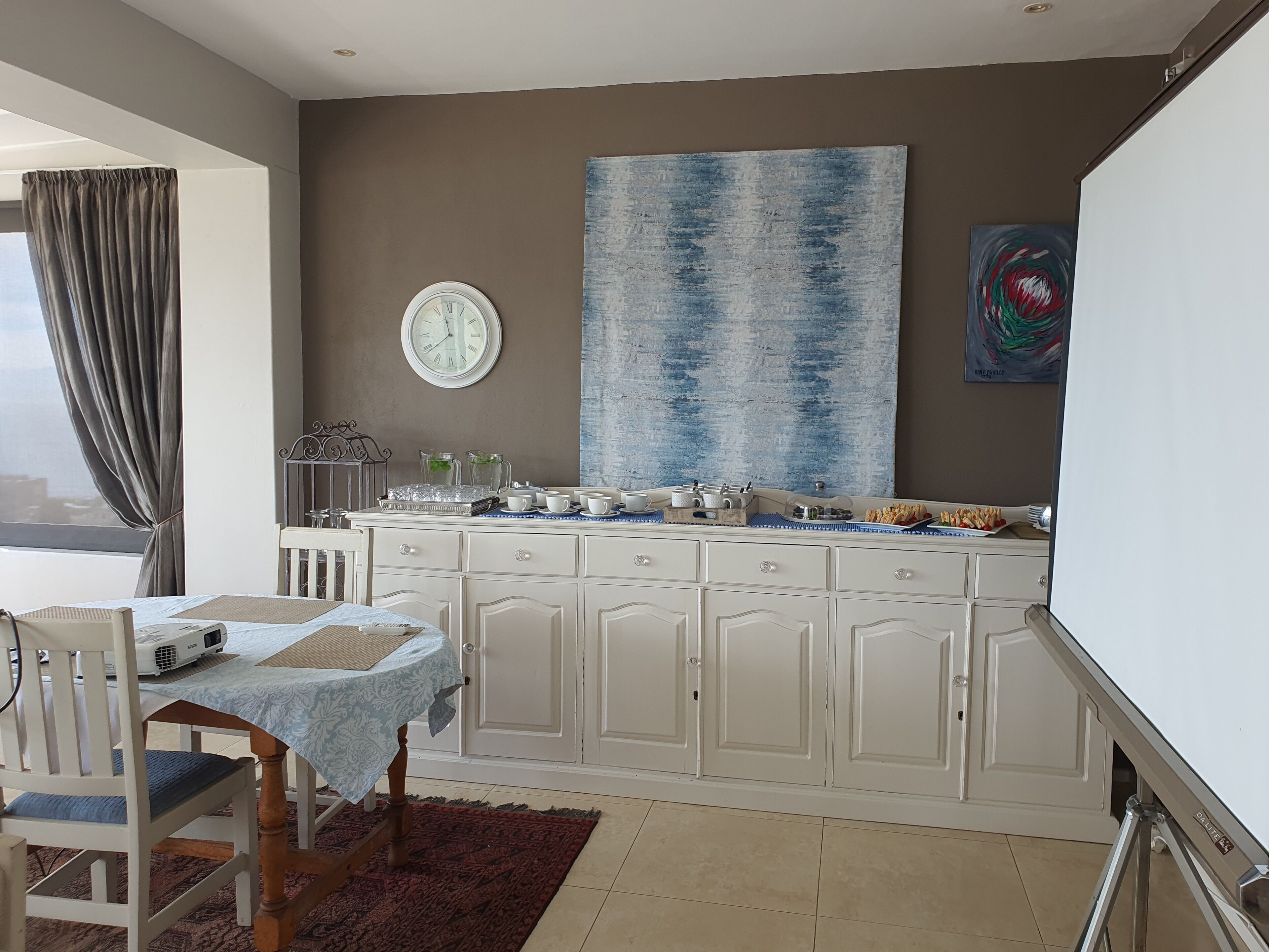 A modern kitchen with a dining area, featuring a large window, a clock, and a blue abstract painting.