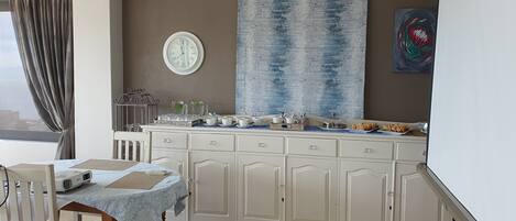 A modern kitchen with a dining area, featuring a large window, a clock, and a blue abstract painting.
