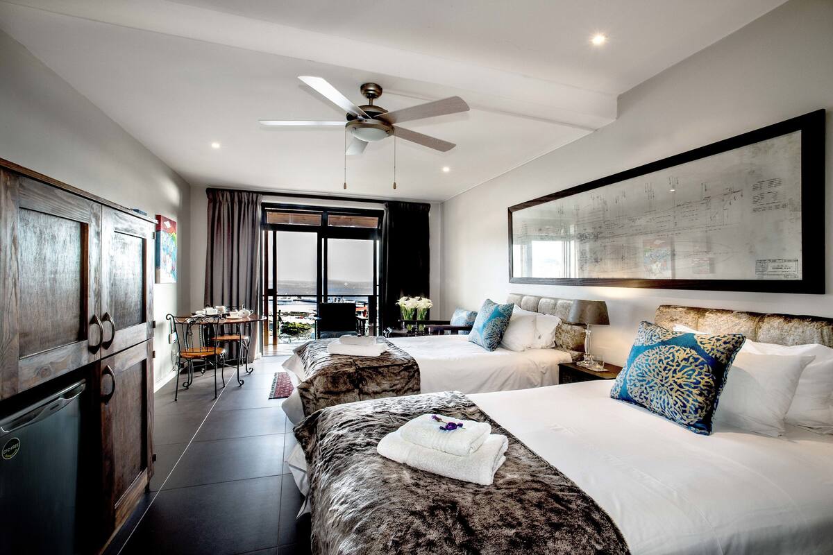 Quarto de hotel com duas camas, área de refeições, ventilador de teto e uma janela grande.
