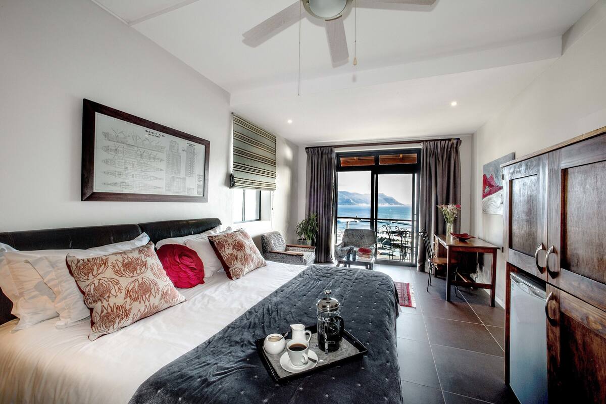 Quarto de hotel com cama, uma mesa de jantar com cadeiras, vista para o mar e um ventilador de teto.